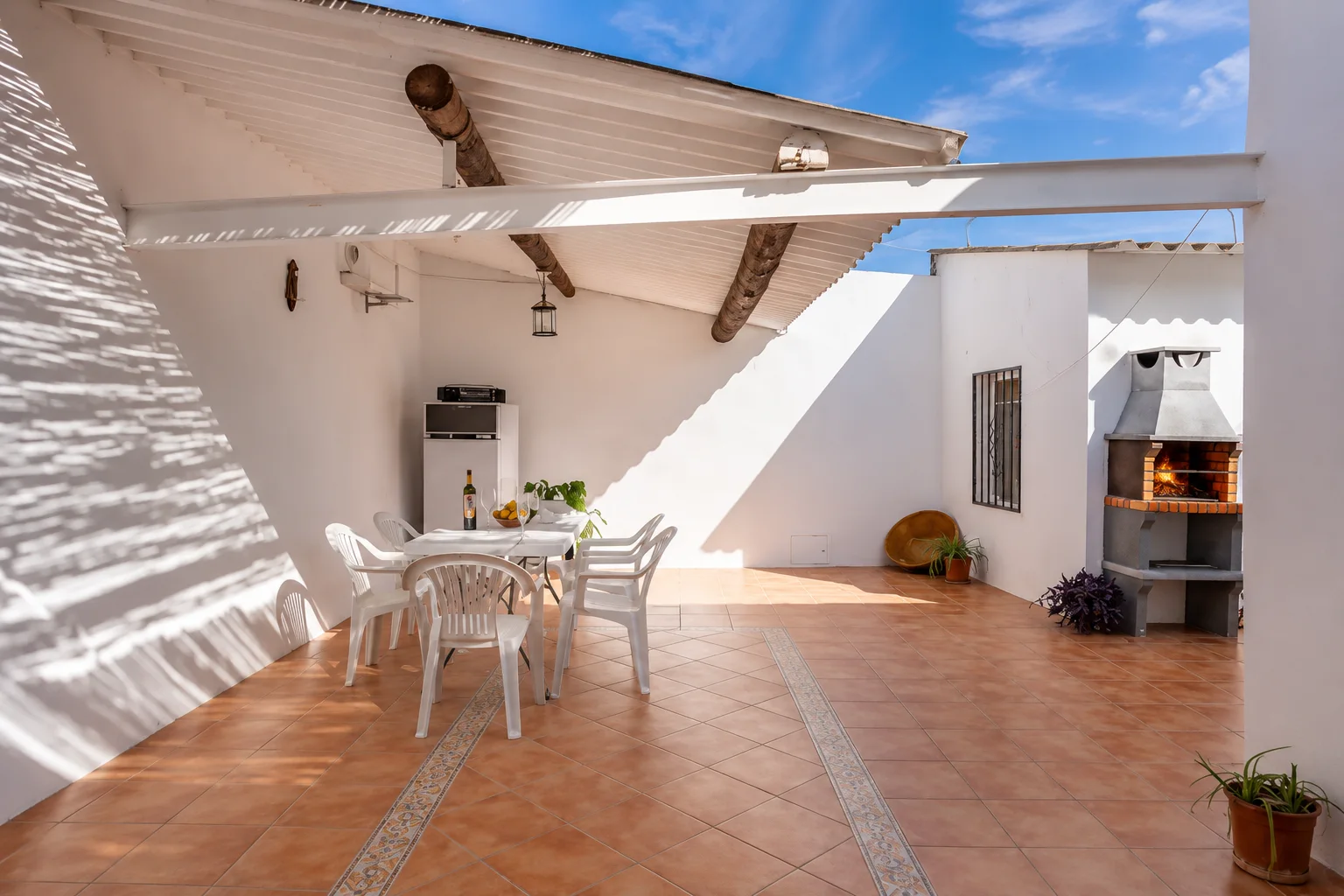 Patio techado con vigas de madera, mesa puesta y la barbacoa de obra al fondo